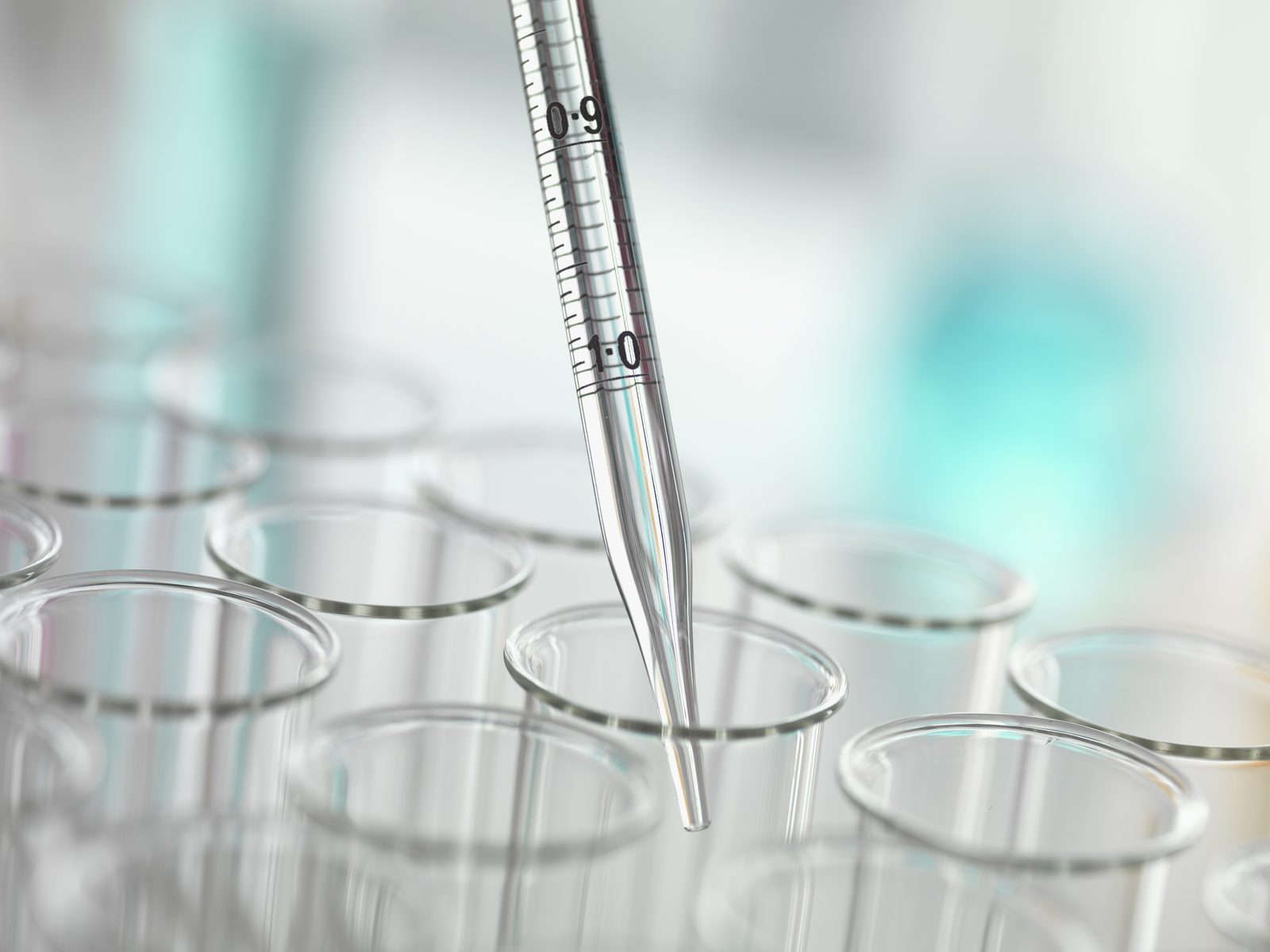 A graduated glass pipette lowered into one of several clear test tubes in a softly lit laboratory with teal tones
