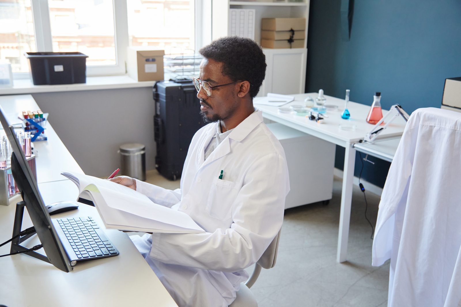 Researcher in a white lab coat and glasses reads a bound record book at a laboratory desk beside a computer and glassware